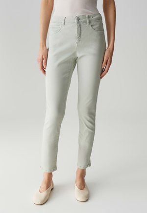 Hellgrüne Jeans mit schmalem Schnitt, Fünf-Taschen-Design und subtilen Steppnähten. Getragen mit beigen, geschlossenen Schuhen.