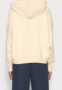 Krämfärgad hoodie med rymlig passform, med en texturerad huva och ribbade muddar. I kombination med mörkblå byxor. Minimalistisk design.