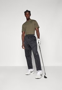 Nike polo shirt in olive green, black golf pants with logo, white sneakers, holding a golf club, wearing a black cap.