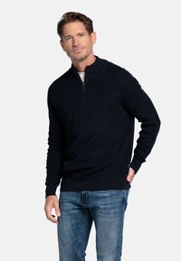 Donkerblauwe gebreide trui met een ritssluiting op de kraag en ribbeltextuur, gecombineerd met lichtblauwe spijkerbroek. Bevat aansluitende mouwen en een zijlabel.