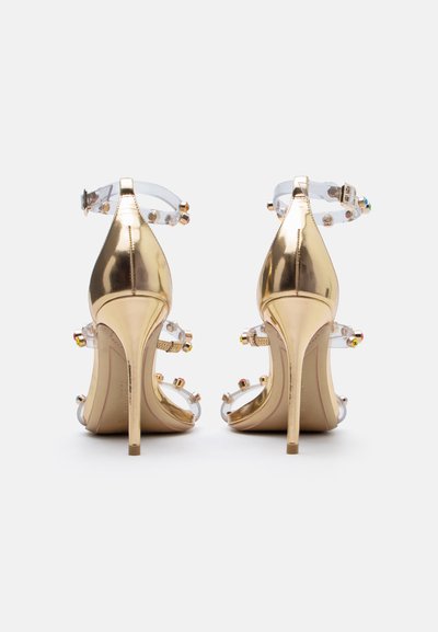Χρυσά σανδάλια στιλέτο με διάφανα λουράκια στον αστράγαλο και τα δάχτυλα, διακοσμημένα με πολύχρωμες πέτρες, απεικονιζόμενα από πίσω.