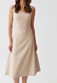 Beige A-linjekjol med en åtsittande bodice och utsvängd kjol. Har en rundad halsringning och slät tygtextur. Utan ärmar.