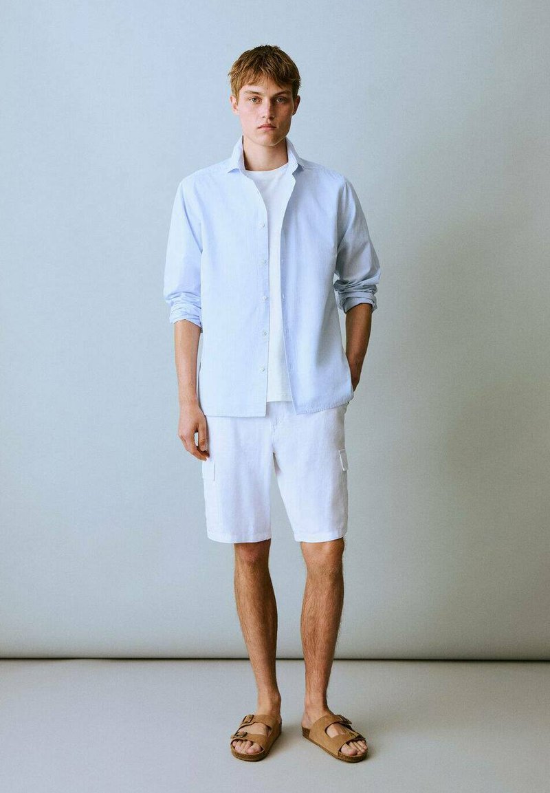 Svetlo modra srajca na gumb, bela majica pod njo, bele kratke hlače iz blaga in rjave sandale. Gladek material in sproščen dizajn.
