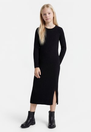 Jeune fille aux longs cheveux blonds portant une robe midi noire côtelée à manches longues avec une fente latérale et des bottines noires sur fond blanc.