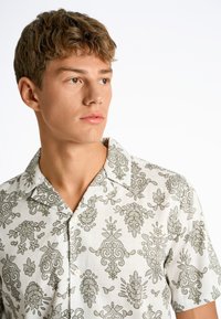 Camisa de manga corta de tela blanca con patrones florales y de cachemira en verde, con cuello acampanado y tapeta de botones.