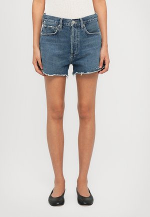 Persoon draagt blauwe denimshorts met versleten zoom en zwarte platte schoenen, staande tegen een effen witte achtergrond.