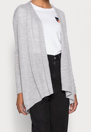 Cardigan grigio chiaro con scollo aperto, maniche lunghe e vestibilità rilassata. Tessuto a maglia con un orlo posteriore leggermente più lungo. Indossato sopra una t-shirt bianca.