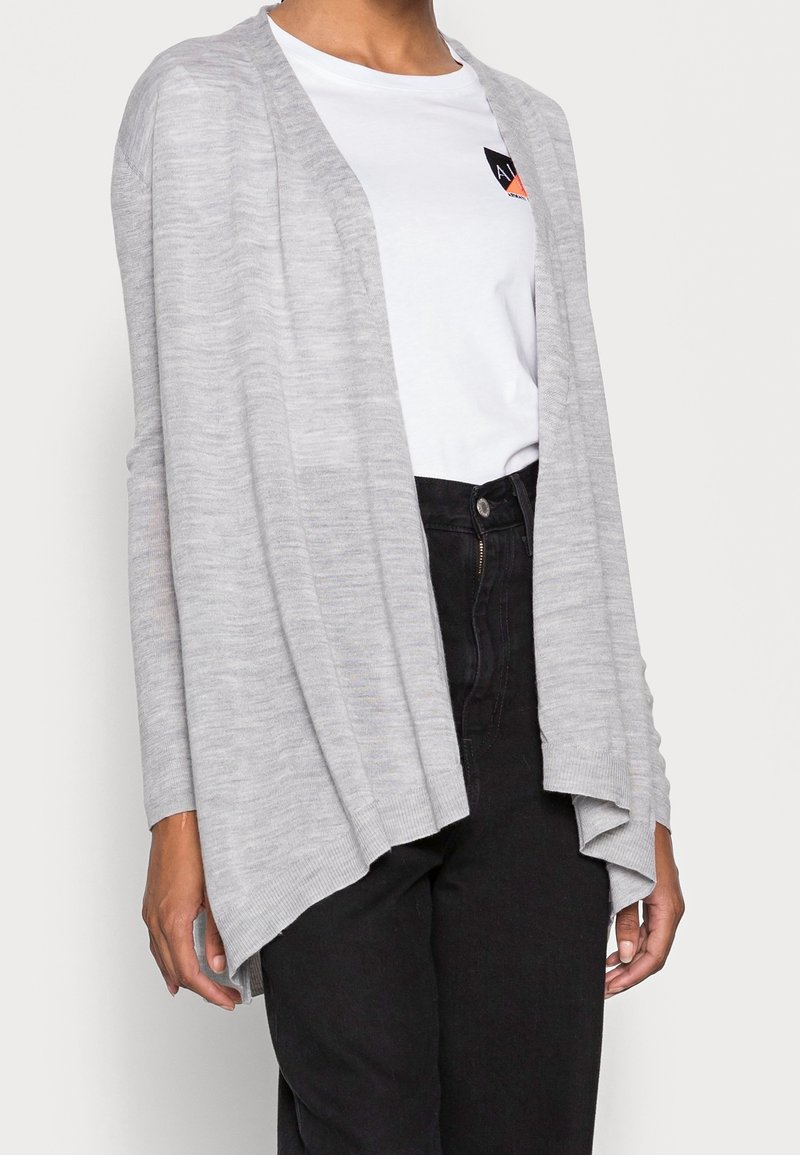 Ljusgrå cardigan med öppen front, långa ärmar och avslappnad passform. Stickad textur med en något längre bakre fåll. Bärs över en vit t-shirt.