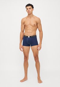 Boxeri navy dintr-un material moale, cu un talie cu logo și un model texturat pe întreaga suprafață. Modelul stă în picioare, desculț.
