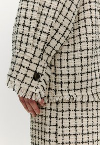 Beige tweed jas met een geruit patroontje in zwart. Bevat rafelige randen en een knoop op de mouw. Structuurstof met hand zichtbaar.