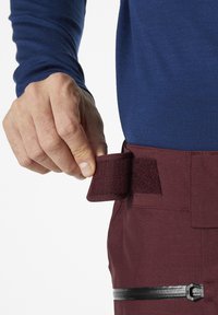 Bordeauxrode broek met een klittenbandsluiting bij de taille, gestructureerde stof, en een zwart ritszakje aan de voorkant. Blauwe langslavende shirt zichtbaar.