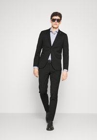 Completo nero su misura con giacca aderente e pantaloni Coordinati. Camicia a quadri blu chiaro sotto. Scarpe nere e occhiali da sole.