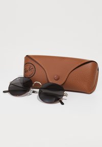 Ochelari de soare Ray-Ban cu lentile negre rotunde, ramă din fibră de broască țestoasă și elemente metalice argintii, îmbinați cu un toc din piele maro texturată.