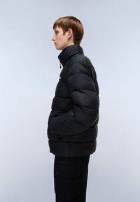 Jachetă puffer neagră cu guler înalt, având secțiuni matlasate și buzunare laterale, expusă pe un model pe un fundal deschis.