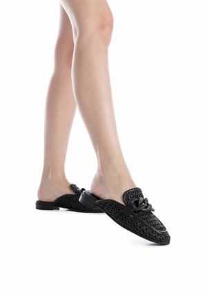 Scarpe slip-on nere con un design intrecciato, caratterizzate da un accento decorativo a nodo e una suola piatta, che evidenziano texture e motivi intricati.