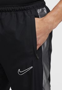 Hand in der Tasche von schwarzen Nike-Hosen mit elastischem Bund und seitlichem grauen Einsatz, versehen mit weißem, gesticktem Nike-Swoosh-Logo.