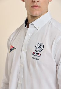Camisa blanca de botón con acentos en el cuello azul, logo bordado del equipo de vela y parches con banderas de colores y nombre de la marca.