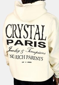Crème hoodie met gedurfde zwarte tekst op de achterkant: "CRYSTAL PARIS Jewelry & Timepieces NO RICH PARENTS," met een relaxte pasvorm en capuchon.
