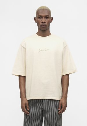 Jeune homme aux cheveux blonds courts porte un T-shirt beige oversize avec le logo "GmbH" et un pantalon rayé noir et blanc, debout sur fond blanc.