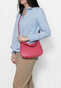 Roze leren handtas met een gebogen vorm en een afneembare schouderband, voorzien van zilveren oogdetails langs de onderrand.