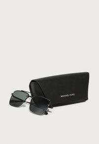 Melnas taisnstūrveida saulesbrilles ar pelēkiem objektīviem, metāla rāmi un plastmasas tempļiem, blakus melnai maciņai ar Michael Kors logo.