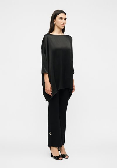 Žena s dugom tamnom kosom koja nosi široku crnu satensku majicu, crne hlače s malim ukrasima i crne cipele otvorenih prstiju, stoji uz bijelu pozadinu.