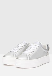 Baskets métalliques argentées avec des lacets blancs, dotées d'une semelle plate et épaisse blanche et d'un petit noeud sur le talon pour une touche d'accent supplémentaire.