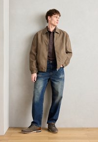 Brun bomberjakke med elastiske mansjetter, en mørk skjorte, falmede blå jeans, og teksturerte grå sko med brune detaljer. Enkelt bakgrunn.