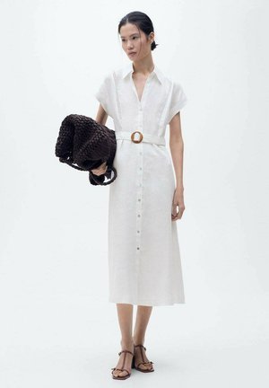 Witte linnen shirtjurk met korte mouwen, knoopsluiting aan de voorkant en een bruine riem met een ronde gesp. Model houdt een donkere gevlochten tote bag vast.