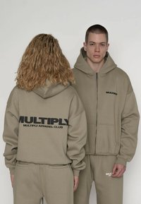 Beżowe bluzy rozpinane z kapturem marki "Multiply Apparel Club" z dużym czarnym logo na plecach, w komplecie z dopasowanymi dresami. Miękki, bawełniany materiał.
