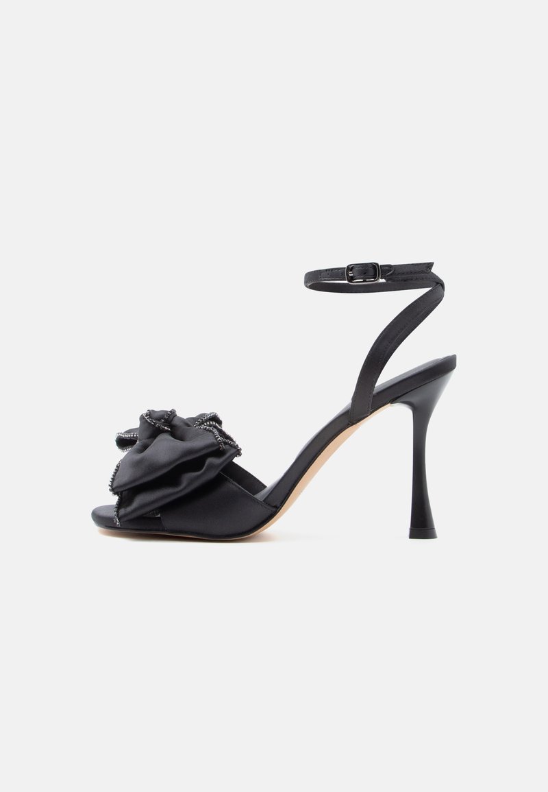 Mustad kontsade mustad sandaalid, millel on pahkluu rihm ja suur rüütud kangast põlledetail varba juures, valge tausta ees.