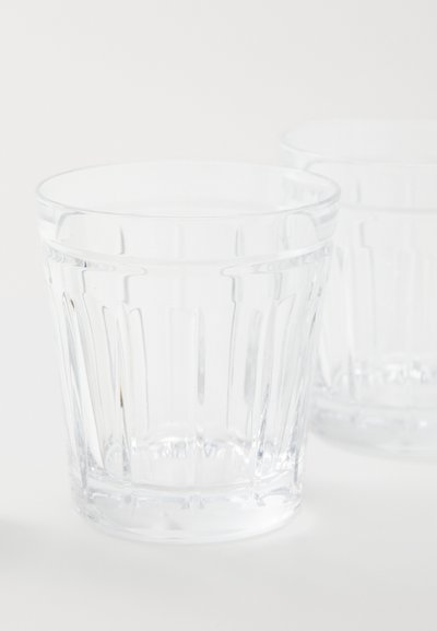 Διάφανα γυάλινα ποτήρια με κατακόρυφη ριγέ υφή, λεία χείλη και στιβαρή βάση; δύο ποτήρια μερικώς ορατά, που αντανακλούν το φως.