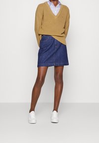 Beige pletený svetr s hlubokým výstřihem do V, kombinovaný s temně modrou džínovou áčkovou sukní. Bíl�é tenisky dotvářejí outfit. Pod ním je vidět pruhovaná košile.