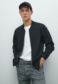 Svart zip-up bomberjacka med kontrasterande grå muddar, bärs över en vit t-shirt. Modellen har matchat med blekta grå jeans och en mörk keps.