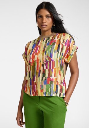 Mujer con cabello largo y negro, que lleva pendientes y pulsera dorados, blusa de manga corta con estampado abstracto multicolor y pantalones verdes, de pie con la mano en el bolsillo.