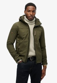 Oliwkowa kurtka softshell z kapturem, z zamykanymi kieszeniami, wysokim kołnierzem i szarym swetrem pod spodem. Ciemne dżinsy i złoty zegarek.