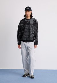 Black oversized sweatshirt with intricate gray graphics, paired with light denim pants and black sneakers featuring yellow accents.