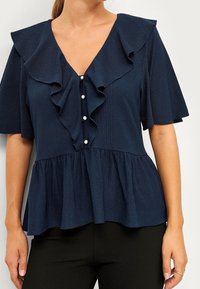 Kvinna iklädd en marinblå blus med volang V-ringning, pärlknappar framtill, lätt puffärmar och peplumslut, i kombination med svarta byxor.