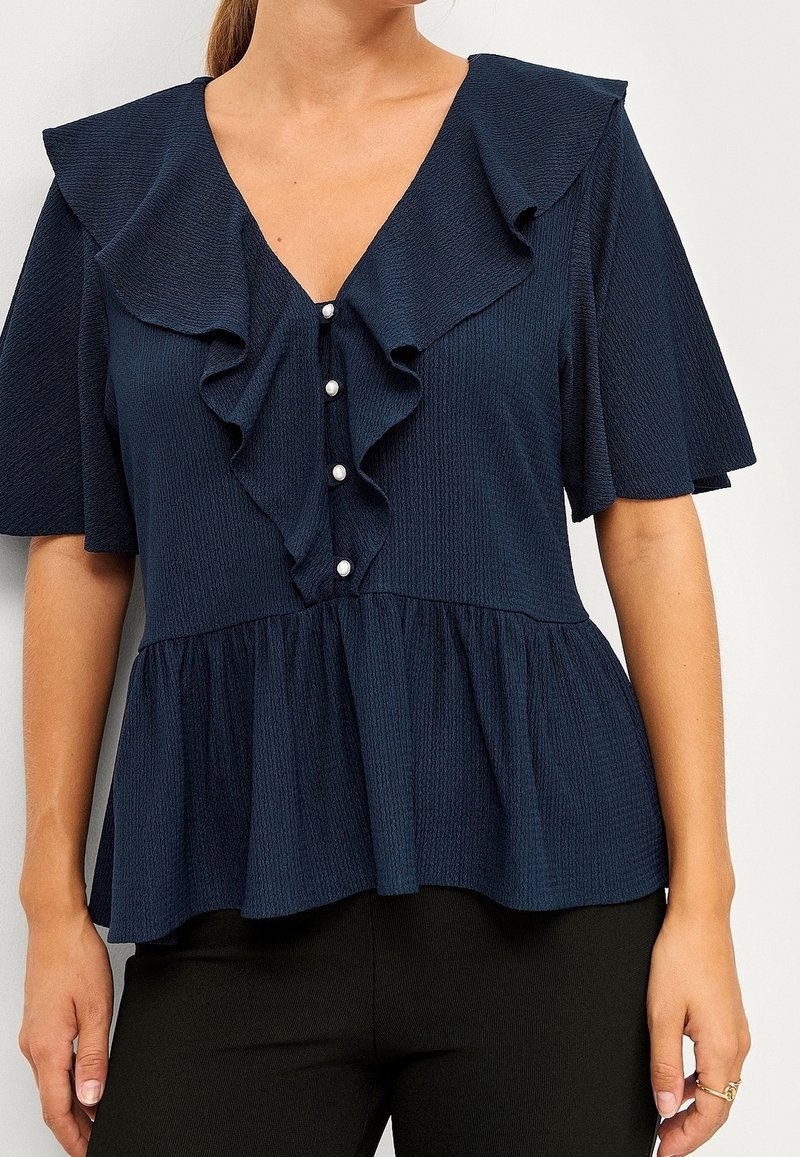 Kvinna iklädd en marinblå blus med volang V-ringning, pärlknappar framtill, lätt puffärmar och peplumslut, i kombination med svarta byxor.