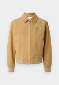 Veste bomber en daim marron avec fermeture zippée, poignets et ourlet côtelés, et un petit logo vert sur la poitrine. Comprend deux poches avant.