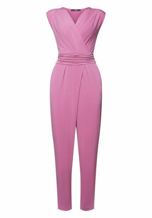 Mujer con vestido cruzado sin mangas de color rosa con cintura fruncida, mano apoyada en la cadera, sobre un fondo azul sólido.