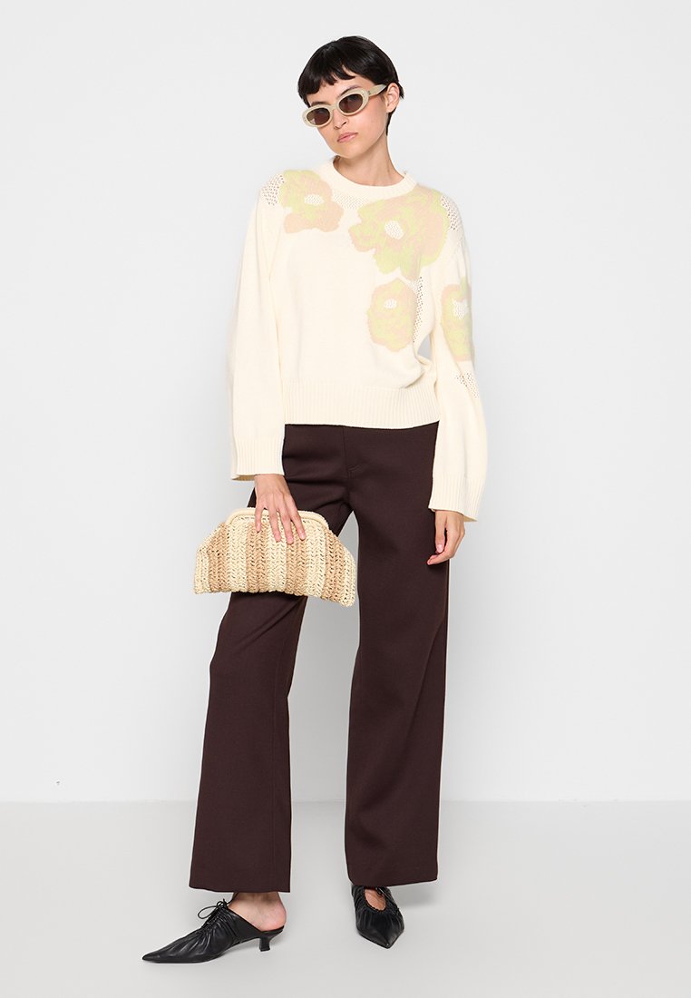 Cremfarbener Strickpullover mit floralen Mustern in Grün und Hellorange, kombiniert mit weit geschnittenen, dunkelbraunen Hosen. Das Model hält eine gewebte beige Tasche.