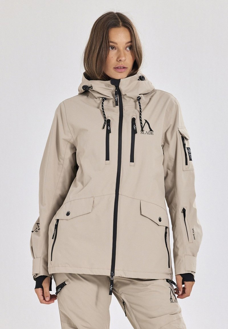 Beige wasserfeste Jacke mit Kapuze, Reißverschluss vorne, mehreren Taschen, Zugbändern und Logodetails. Glatte Textur, entworfen für den Einsatz im Freien.