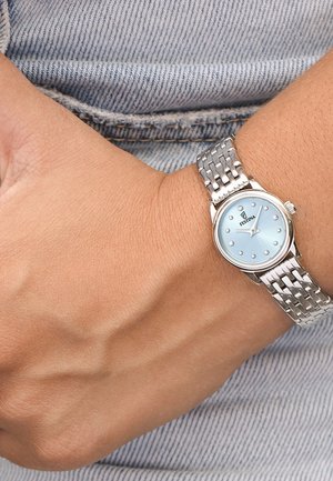 Polso con orologio a cinturino metallico argentato che mostra le 3:12, quadrante azzurro chiaro e sfondo in tessuto di jeans.