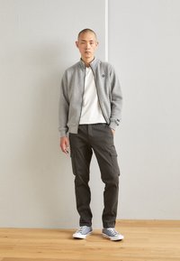Gray quilted bomber jacket with a zip front, worn over a white shirt, paired with dark cargo pants and gray sneakers. Minimalist design.