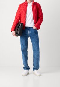 Giacca rossa trapuntata, polo bianca, jeans blu e sneakers bianche. Borsa nera tenuta in un mano, in piedi contro uno sfondo neutro.