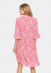 Blommig klänning i rosa tyg med vita blommönster, med en design av utsvängda ärmar och knälång fåll. Baksidan visas.