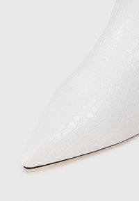 Zapato blanco de punta afilada con textura brillante de cocodrilo embossado y delgada línea negra en la suela, sobre un fondo blanco liso.