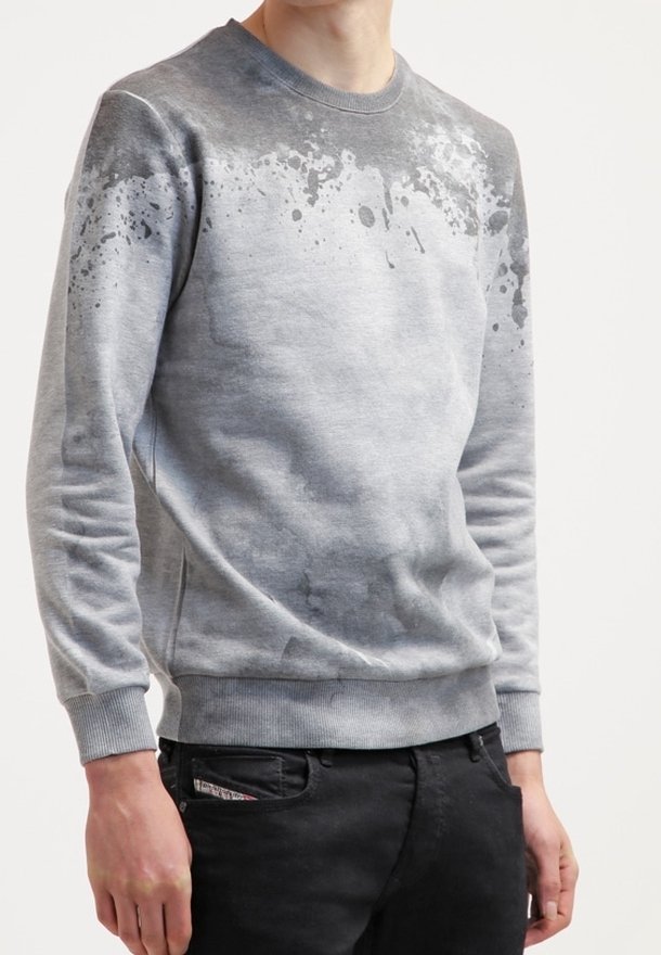 Homme portant un sweat-shirt gris clair avec un motif d'éclaboussures de peinture gris foncé sur le haut de la poitrine et les épaules, associé à un pantalon noir.