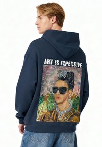 Felpa con cappuccio blu navy con una grande grafica di Frida Kahlo sulla schiena, circondata da fogliame vivace e la scritta "L'ARTE È COSTOSA."